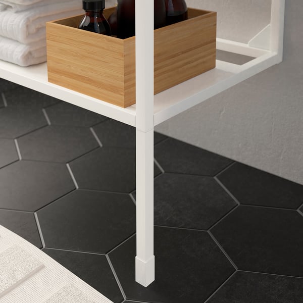 Pieds d’étagère ENHET blancs réglables, contenant une boîte de rangement en bois avec des bouteilles, posée sur des carreaux de sol hexagonaux foncés.