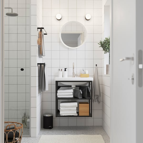 Salle de bain minimaliste : carrelage blanc, miroir rond, meuble-lavabo ENHET avec serviettes, boîte en bois, douche en verre, pomme de pluie, chaise en bois avec des vêtements.