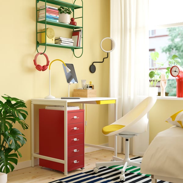 Bureau aux murs jaunes, bureau rouge, chaise blanche, ordinateur portable, plante, tapis, étagère verte avec des livres, et une fenêtre.