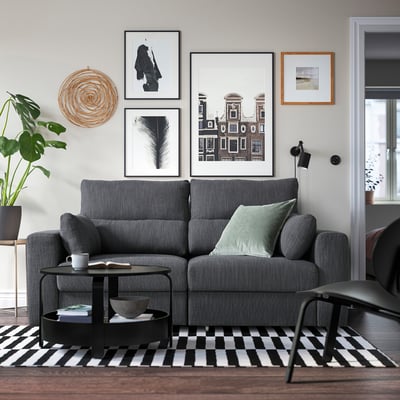 Salon moderne avec grand canapé ESKILSTUNA gris, table basse noire, tapis noir et blanc, art mural et plantes.