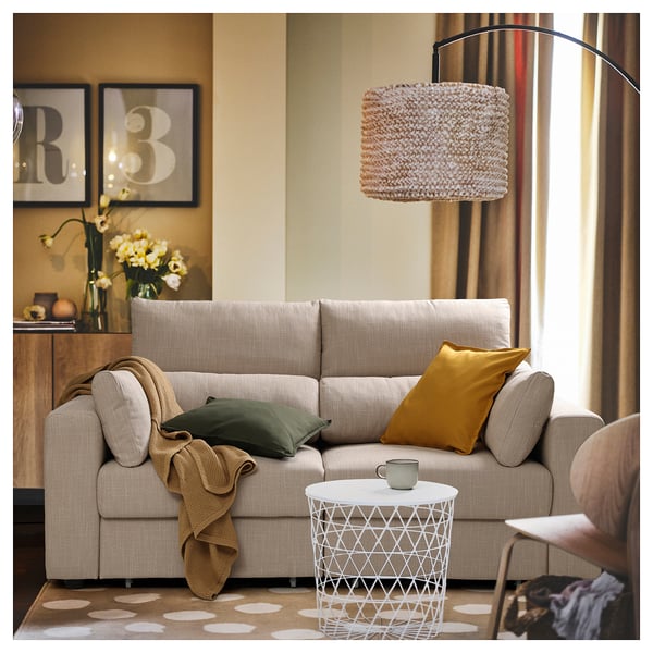 Salon confortable avec canapé ESKILSTUNA beige, coussin moutarde, lampe tressée et table d’appoint avec tasse.