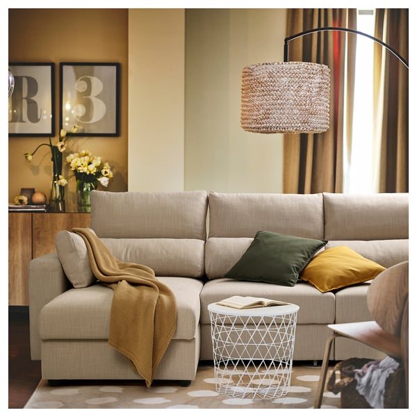 Sectionnel ESKILSTUNA beige avec appuie-tête réglables, chaise longue amovible, coussins verts et moutarde, sous une suspension.