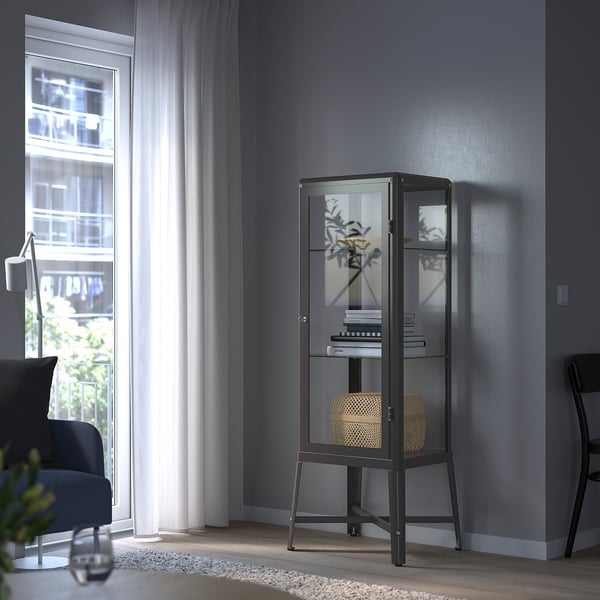 Armoire à porte vitrée noire dans un coin, exposant livres, vase et panier.