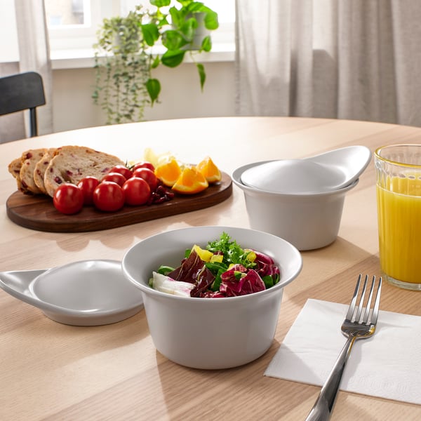 Table à manger en bois avec bols, pain, fruits et jus. Le bol central a une salade colorée, entourée de fourchettes, de serviettes et de bols blancs couverts.