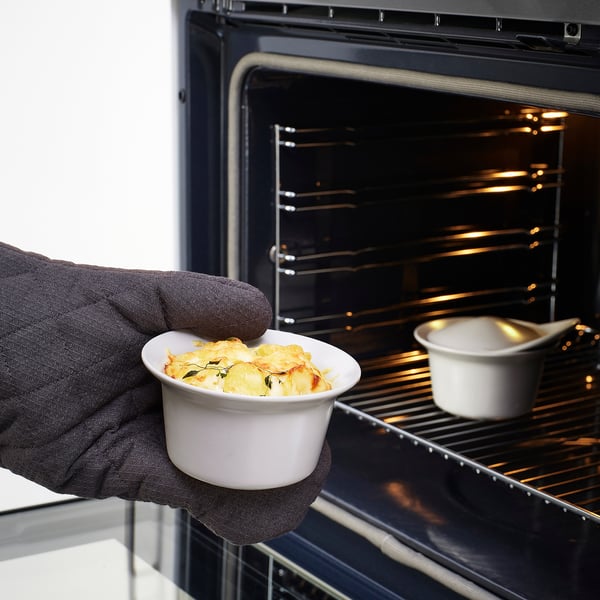 Portant un gant de cuisine tenant un plat blanc allant au four avec des aliments cuits, prêt à être placé dans un four noir avec un autre plat à l’intérieur.