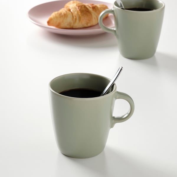 Deux tasses vertes avec des cuillères, l’une remplie de café, à côté d’une assiette avec un croissant.