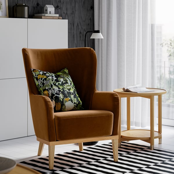 Pièce lumineuse avec chaise FAGERBACKEN, velours marron, dossier haut, à côté d’une petite table d’appoint en bois sur tapis rayé, à côté d’une grande fenêtre.