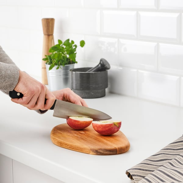Personne tranche une pomme sur une planche à découper en bois d’acacia FASCINERA ; Comptoir de cuisine, dosseret de carreaux de métro blancs visibles.
