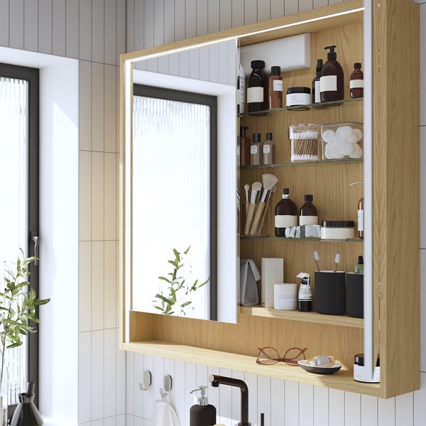 Armoire de toilette en bois avec éclairage intégré, contenant divers articles de toilette et cosmétiques, montée au-dessus d’un évier.