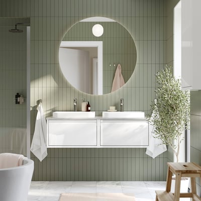 Salle de bain moderne avec miroir rond rétroéclairé FAXÄLVEN. Carreaux vert tendre, vanité blanche et décor végétal. Taille du miroir remarquable, plus grand que le tabouret de tortue.