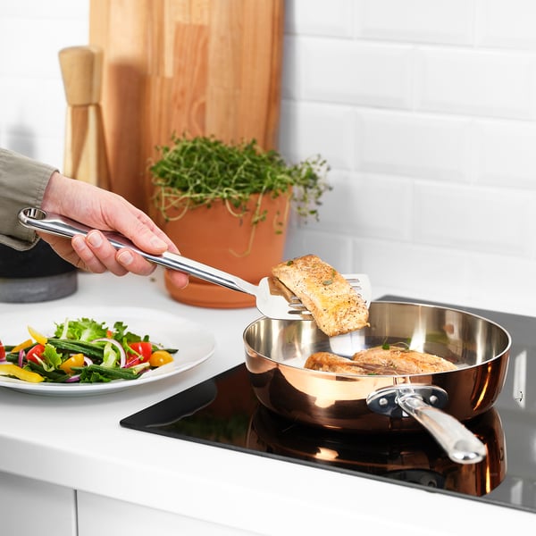 Une main utilise la spatule FINMAT pour retourner le poisson dans une poêle, montrant ainsi sa capacité à glisser sous les aliments et à reposer sur le bord de la poêle.