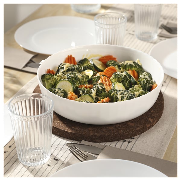Un bol blanc de légumes vapeur repose sur une table en bois, accompagné de tasses et d’assiettes en verre transparent.