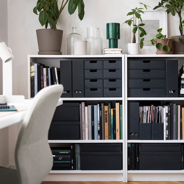 Étagère moderne avec des classeurs de magazines noirs, des livres, des plantes et des vases. Chaise partiellement visible.