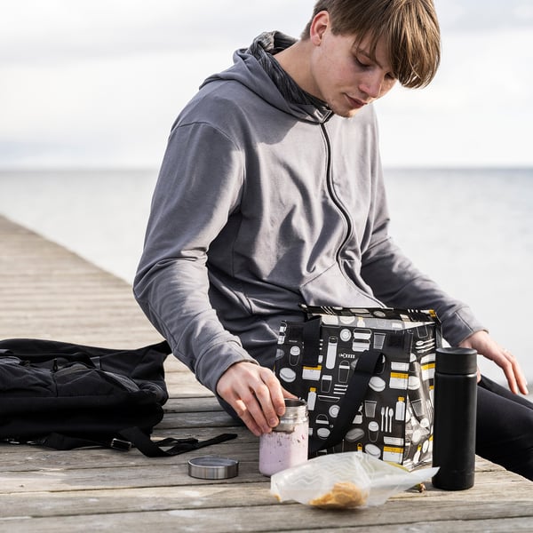 Un homme est assis sur le quai, tenant un sac à lunch FLADDRIG gris avec un motif sur le thème du café. À côté de lui, un sac à dos noir et une bouteille sont placés.