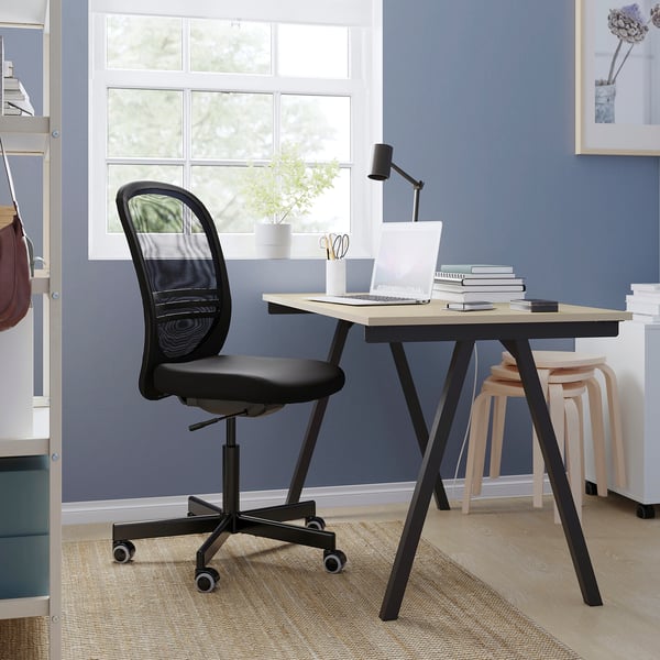 Configuration du bureau à domicile avec une chaise ergonomique en maille noire, un bureau en bois, un ordinateur portable, une lampe et un tabouret contre un mur bleu.