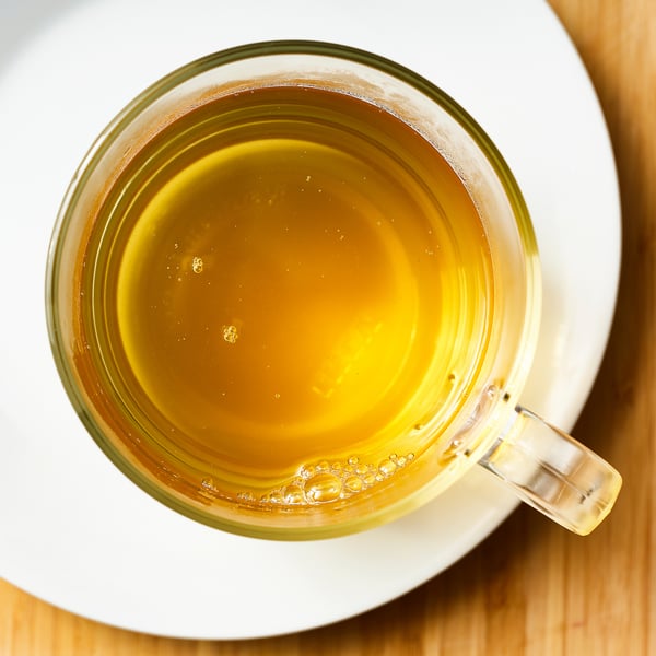 Tasse en verre transparent remplie de thé vert fumant sur une soucoupe blanche.