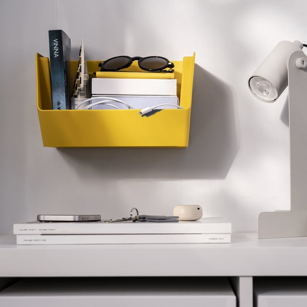 L’organisateur mural jaune vif peut contenir des livres, des lunettes de soleil et des câbles. À côté d’une table avec une lampe.