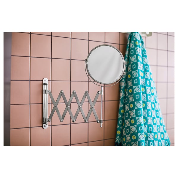 Un miroir loupe FRÄCK argenté monté sur un mur carrelé rose à côté d’un rideau de douche à motifs bleus.