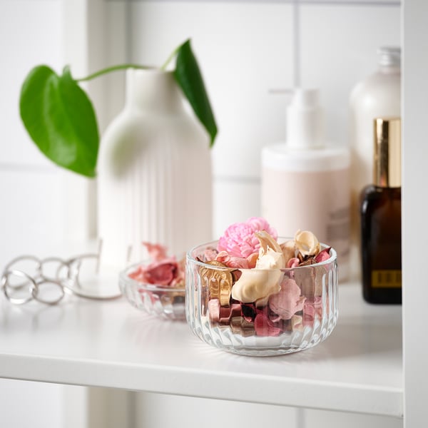 Un petit bol en verre rempli de pot-pourri coloré se trouve sur une étagère blanche. À côté d’un vase blanc avec une plante verte et des produits de beauté.