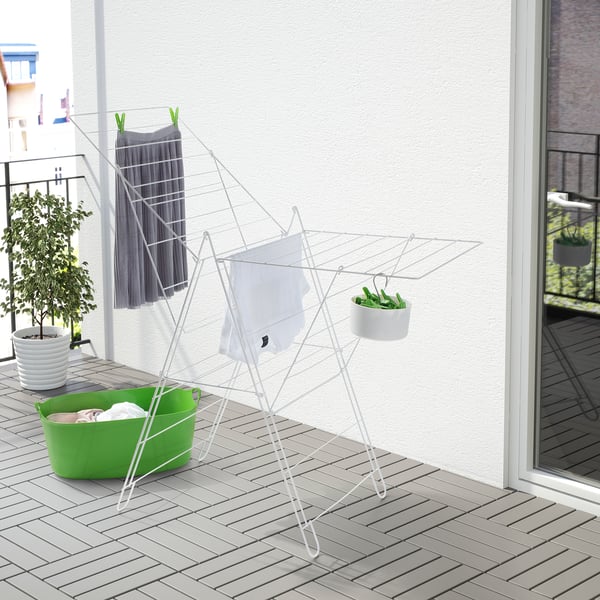 Séchoir FROST blanc sur le balcon, plus grand que le panier à linge à proximité, contient des vêtements et des plantes suspendues.