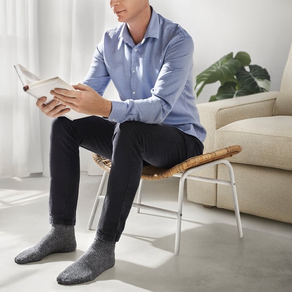 Homme assis sur un tabouret tissé, livre de lecture. Chambre lumineuse avec canapé, plante et fenêtre.