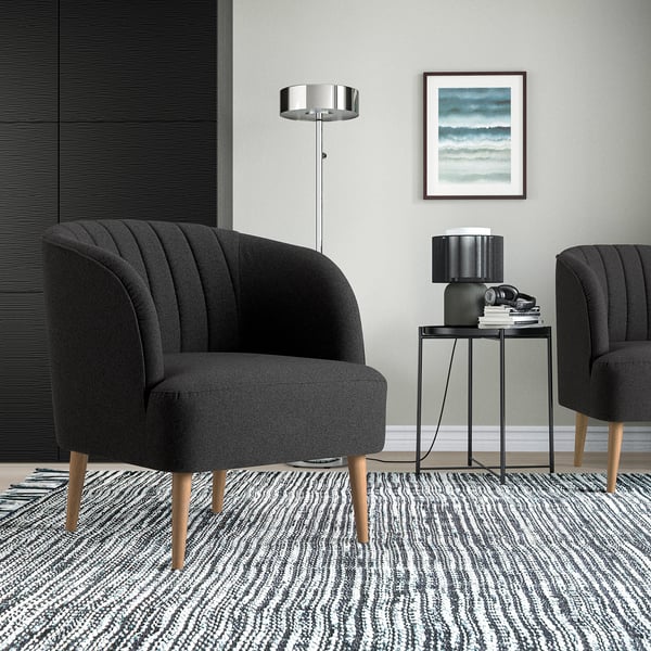 Salon moderne avec deux chaises tonneau FULLÖSA grises sur un tapis rayé, table d’appoint noire avec livres et lampe, mur gris avec art.