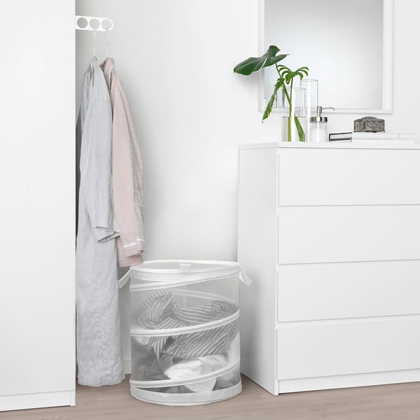 Panier à linge FYLLEN blanc entre la commode et le portemanteau avec des vêtements pliés.