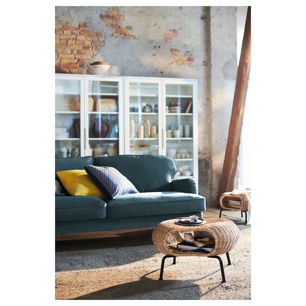 Tabouret GAMLEHULT bleu, rotin tressé, de forme ronde, design rustique, à côté d’un canapé vert, près d’un mur de briques, sous un éclairage chaleureux.