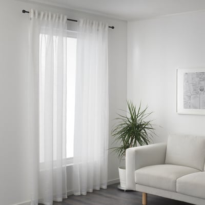 Rideaux transparents près de la fenêtre, la lumière du jour filtre. Fauteuil et plante côté droit.