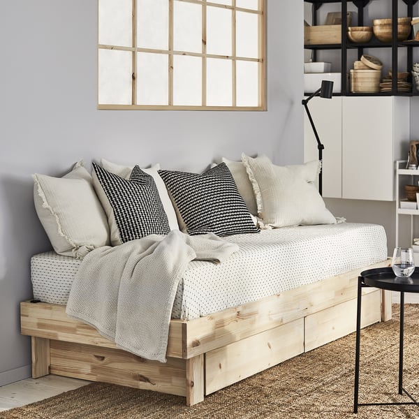 Méridienne moderne en bois clair avec 3 oreillers blancs et 2 oreillers noirs, couverture grise, sous lampe noire.