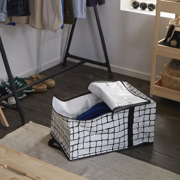 Un sac de rangement GÖRSNYGG sur un sol en bois, motif de grille gris-noir, partiellement rempli de vêtements, près d’un porte-chaussures.
