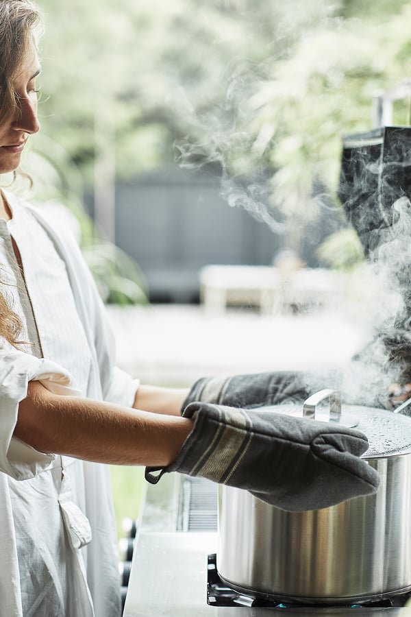 Femme cuisinant à l’extérieur à l’aide d’un gril GRILLSKÄR blanc avec de la fumée montante, portant des gants de cuisine gris, sur un fond de jardin vert.