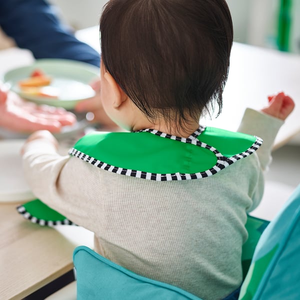 Un enfant vêtu d’un bavoir vert imperméable avec une bordure à carreaux est assis dans une chaise haute et mange. Le bavoir est réglable et facile à nettoyer.