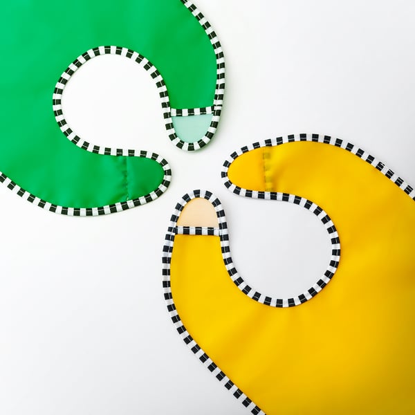 Trois bavoirs aux couleurs vives, vert, beige et jaune, posés à plat, bordés de bordures à carreaux noirs et blancs.