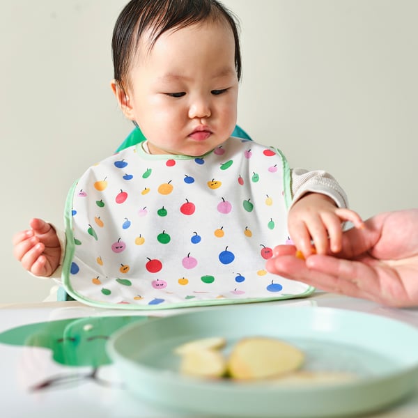 Bébé portant un bavoir coloré avec des poches, assis dans une chaise haute en train de manger. Le bavoir attrape la nourriture, gardant les vêtements propres.