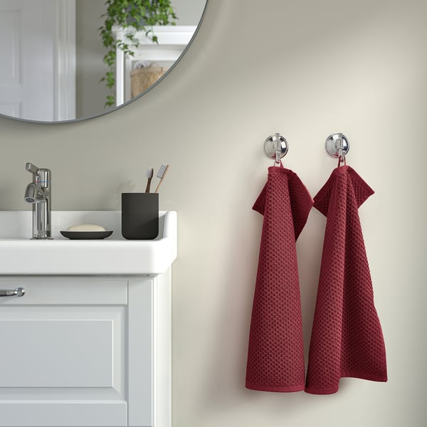 Des serviettes rouges GULVIAL sont accrochées à des crochets muraux à côté d’un évier blanc et d’un porte-brosse à dents noir.