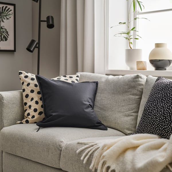 Salon moderne avec GURLI, un coussin en coton noir, sur un canapé gris, visuellement complété par des oreillers à motifs et une couverture douillette.