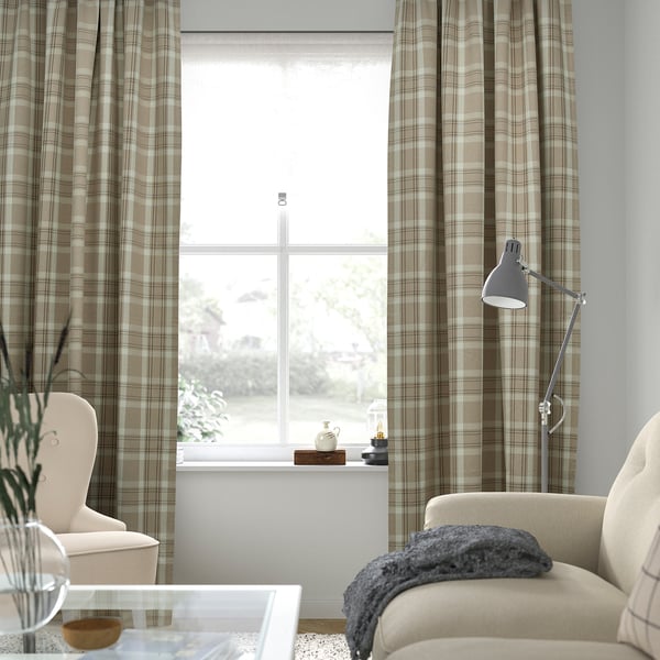 Salon confortable avec de longs rideaux beiges HÄGGVECKMAL. La chaise, la lampe et la couverture ajoutent du confort. Vue de la fenêtre à l’extérieur.