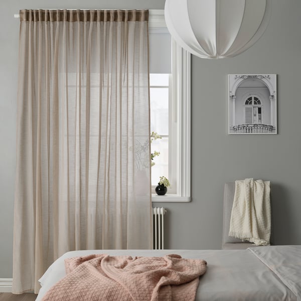 Chambre douillette aux murs gris. Grand lit, couvertures roses, rideaux beiges transparents. Lumière suspendue, plante de fenêtre, oreiller gris.