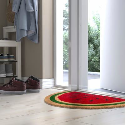 Paillasson rouge vif en forme de pastèque avec écorce verte sous un porte-chaussures, près d’un manteau bleu et d’une porte.