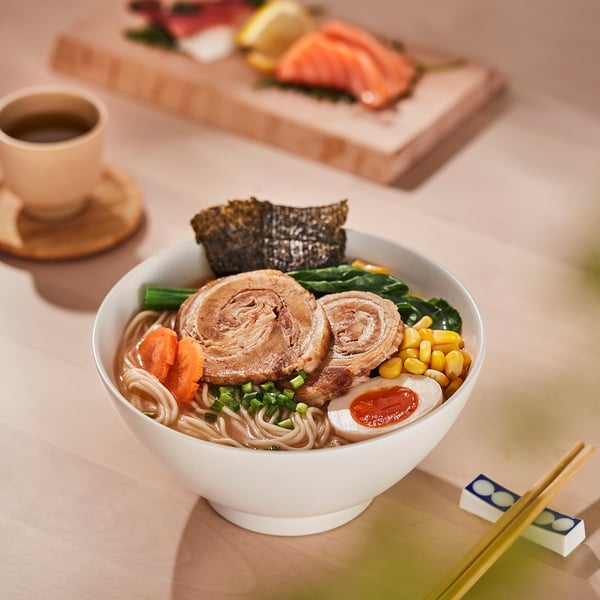 Un bol de nouilles ramen avec des légumes et de la viande ; des baguettes et du thé à côté.
