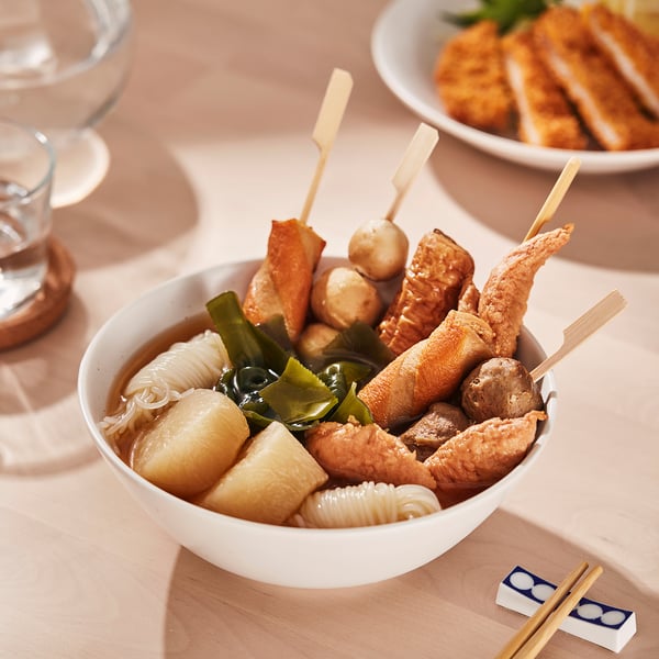 Bol en porcelaine blanche rempli de brochettes d’aliments, de nouilles et de légumes ; Exposé sur une table avec des baguettes.
