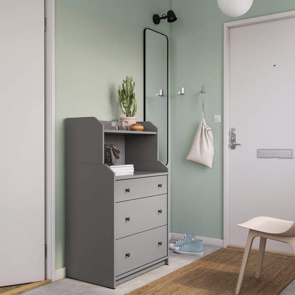 Une commode HAUGA grise avec 3 tiroirs et une étagère supérieure contre un mur végétal. Il comprend un miroir, une plante, une lumière et un porte-chaussures.