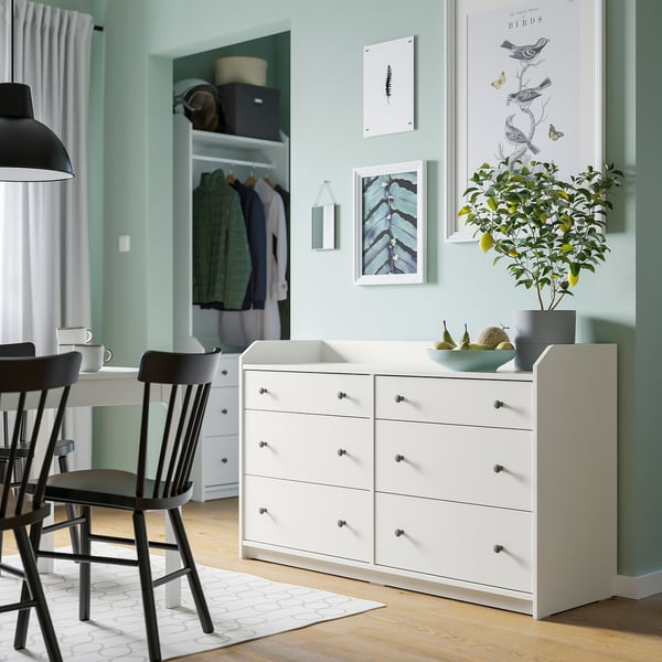 Chambre moderne avec des murs verts, des tiroirs HAUGA, une corbeille de fruits, des plantes, des œuvres d’art, des chaises noires et une suspension.