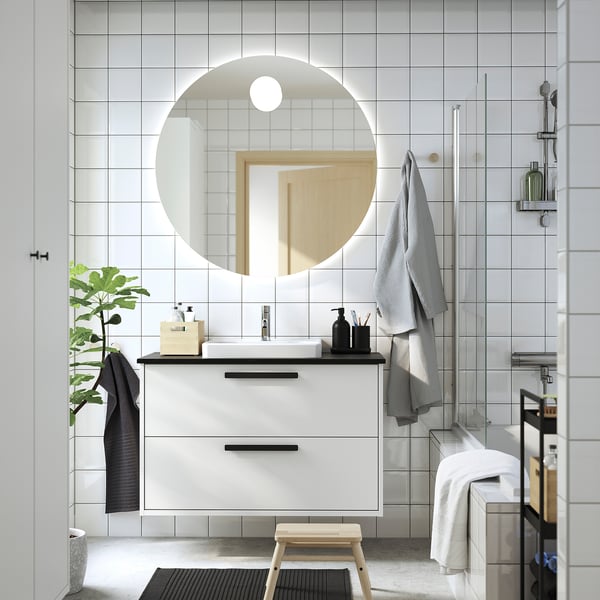 Salle de bain blanche moderne avec coiffeuse HAVBÄCK , miroir rond, accents noirs, serviettes grises et tabouret en bois.