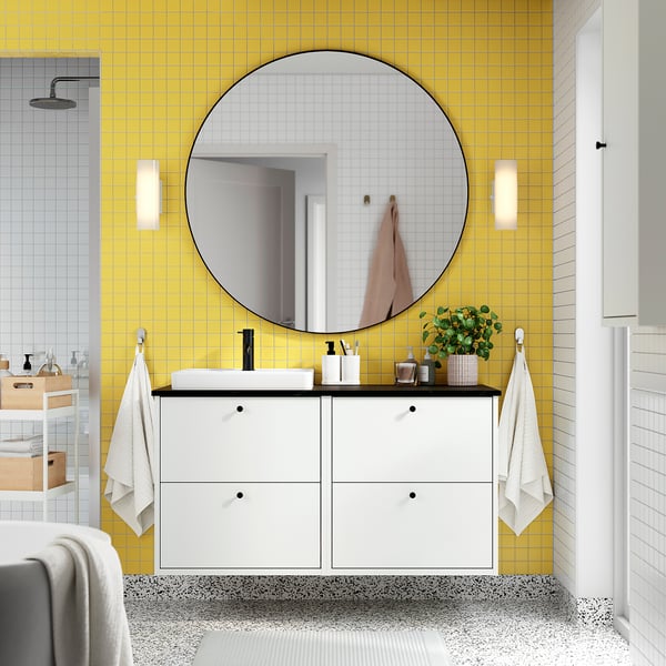 Salle de bain moderne avec murs carrelés jaunes, grand miroir rond, meuble de rangement blanc, lavabo rectangulaire, petites plantes et crochets à serviettes fixés au mur.