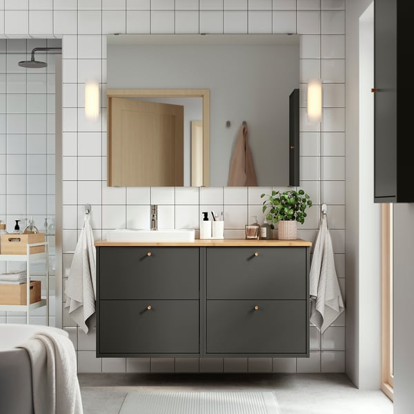 Salle de bain moderne avec vasque gris foncé, plan de travail en bambou, lavabo blanc. Des poignées murales en or. Miroir avec lumières, serviettes, plante.
