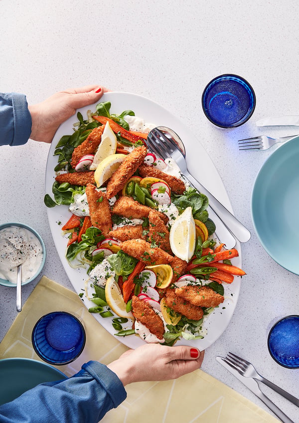 Un plateau de bouchées de poisson panées croustillantes servies avec une salade. Les mains se tendent pour servir. Verres et argenterie bleus.