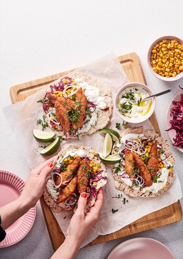 Deux tacos au poisson croustillants sur une planche en bois, garnis de maïs, de citron vert et d’herbes. Une main tient un taco, avec des garnitures et des assiettes supplémentaires à proximité.