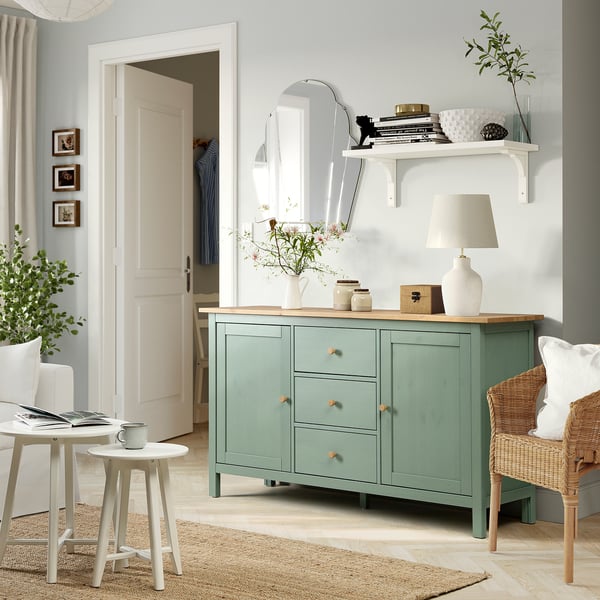 Buffet HEMNES gris-vert avec tiroirs et décor en pin. Chambre confortable avec table pour ordinateur portable et chaise en osier, placard en arrière-plan.